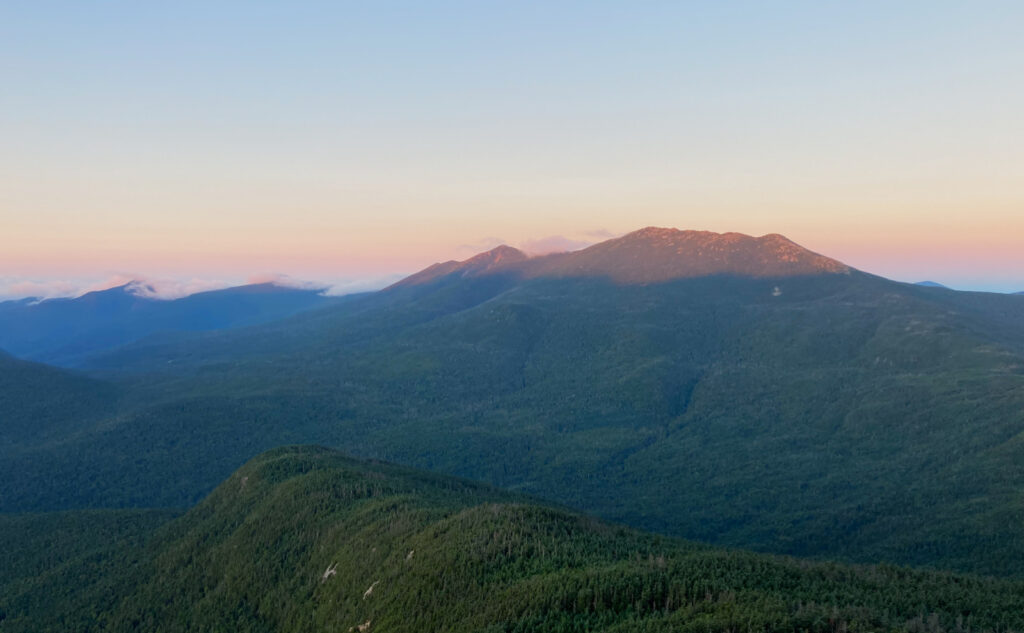 Sunrise dappling the summits of Little Haystack Mountain, Mount Lincoln, and Mount Lafayette. Clouds obscure Mount Liberty and Flume. 